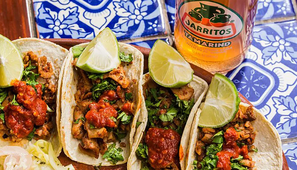 The image shows three flavorful tacos garnished with lime on a colorful patterned surface accompanied by a bottle of Jarritos Tamarind soda
