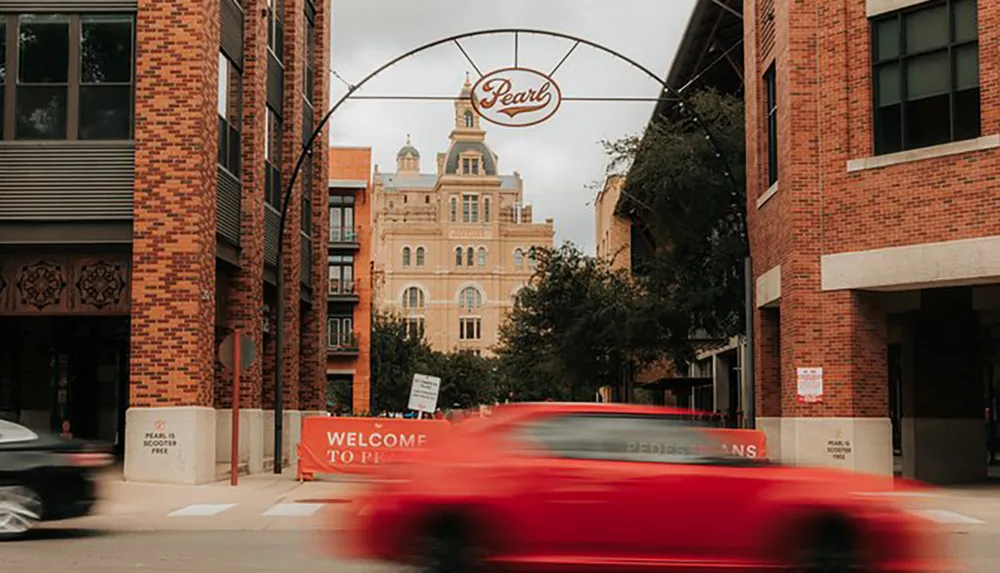 A blurred red car speeds past urban brick buildings under the Pearl archway on a cloudy day