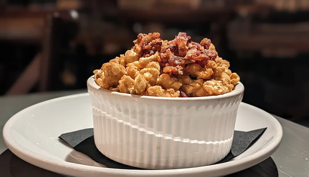 A bowl filled with popcorn topped with crispy bacon pieces is presented on a white plate with a dark napkin underneath