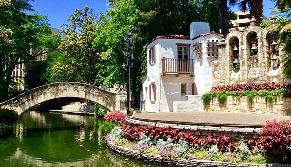 The image shows a picturesque scene with a quaint white building next to a calm river a stone bridge and vibrant flowering plants under a bright blue sky