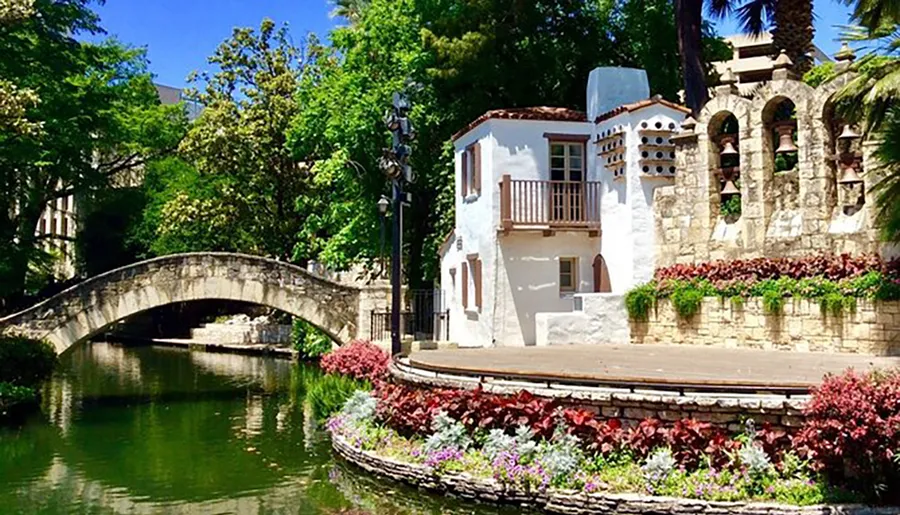 The image shows a picturesque scene with a quaint white building next to a calm river, a stone bridge, and vibrant flowering plants under a bright blue sky.
