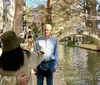 A couple is taking a selfie by a canal with a stone bridge in the background while an older man stands in the foreground looking towards the camera