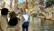 A couple is taking a selfie by a canal with a stone bridge in the background, while an older man stands in the foreground looking towards the camera.