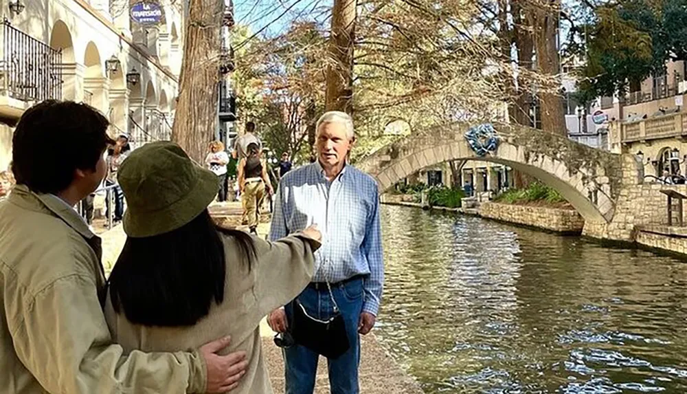 A couple is taking a selfie by a canal with a stone bridge in the background while an older man stands in the foreground looking towards the camera