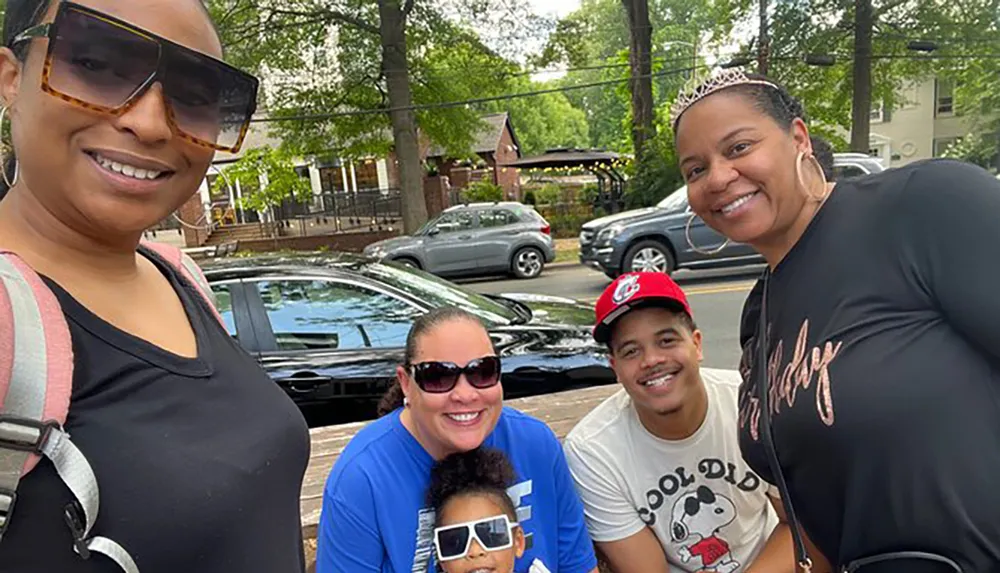 A group of five people including a child are happily posing for a selfie outdoors on a sunny day