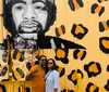 A couple is posing for a photo in front of a vibrant yellow wall featuring an artistic mural with black and orange leopard print and a large portrait