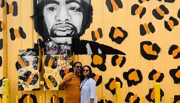 A couple is posing for a photo in front of a vibrant yellow wall featuring an artistic mural with black and orange leopard print and a large portrait.