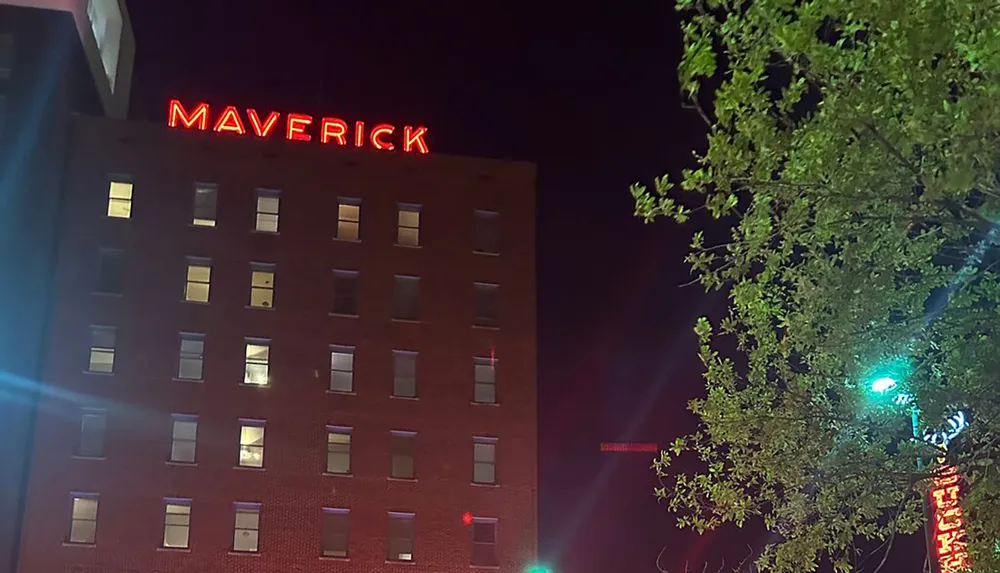 The image shows a nighttime view of a building with the red neon sign MAVERICK at the top some illuminated windows and the silhouette of a tree in the foreground