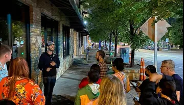 A group of attentive people listen to a speaker during an evening gathering on a city sidewalk lined with trees and storefronts.