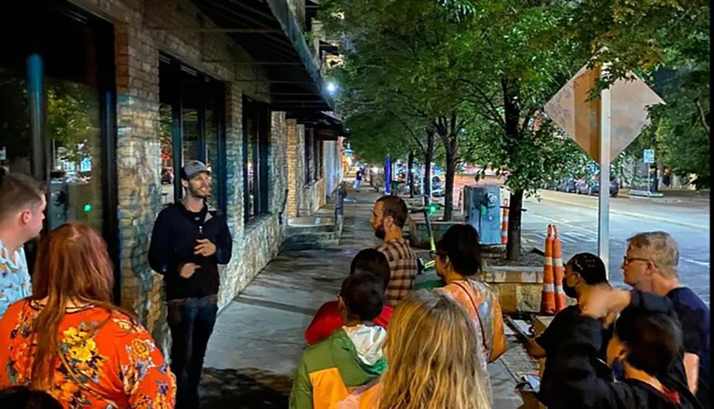 A group of attentive people listen to a speaker during an evening gathering on a city sidewalk lined with trees and storefronts