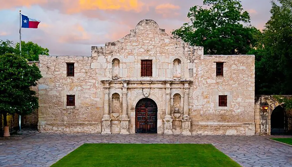 The image shows the historic Alamo mission in San Antonio Texas under a cloudy sky with the Texas state flag flying nearby