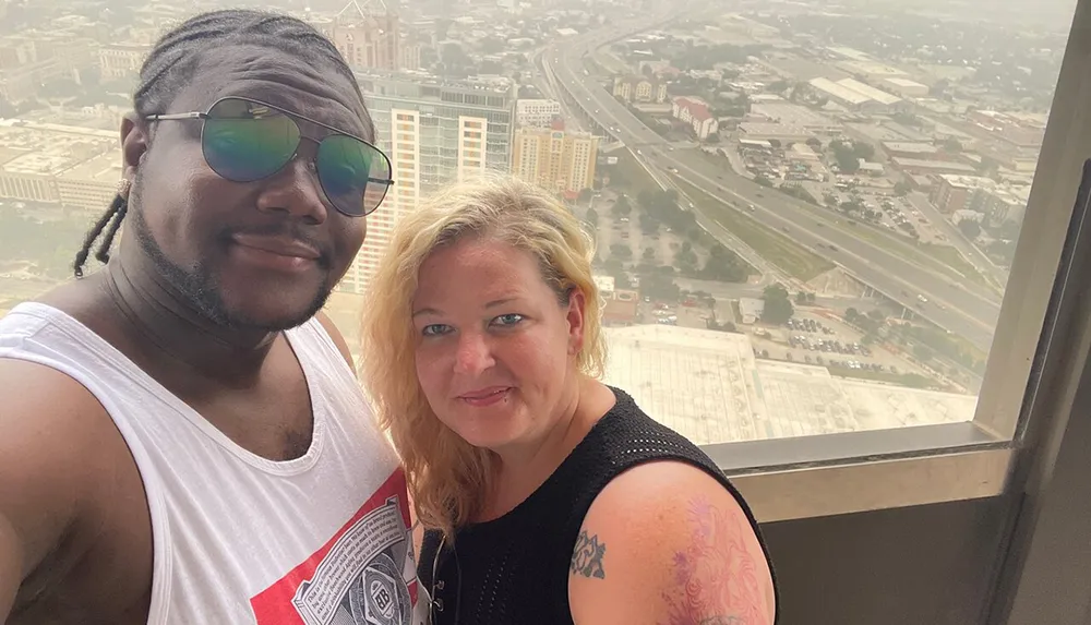 A man and a woman are posing for a selfie with a hazy city skyline in the background