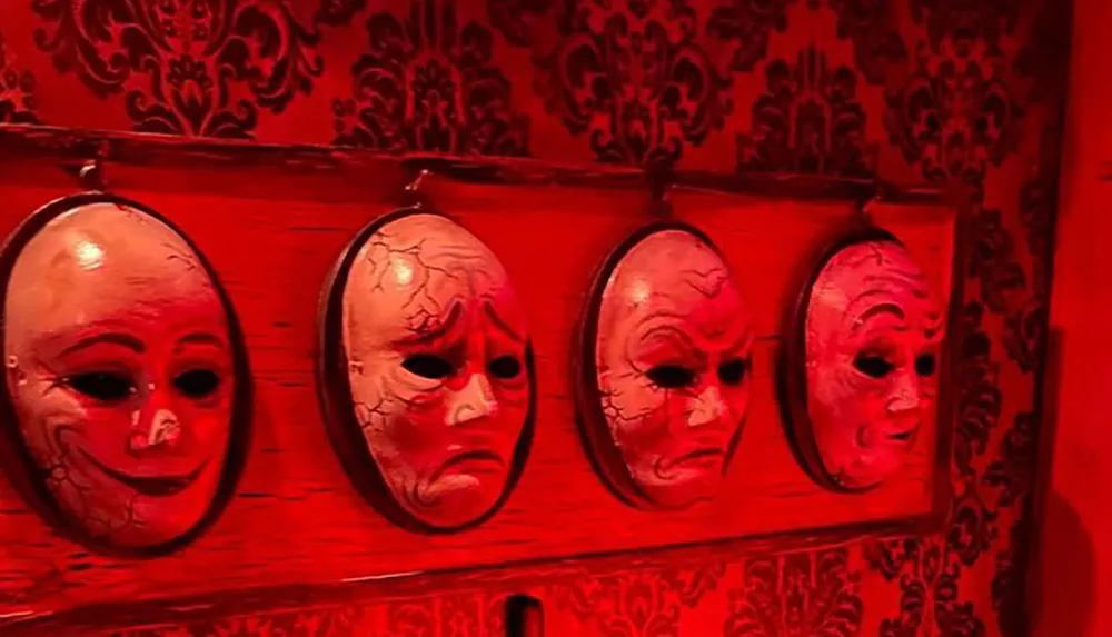 Four theatrical masks exhibiting a range of emotions from neutral to distressed hang on a red wall with ornate patterns