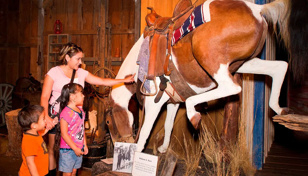 A woman and two children are interacting with a taxidermied horse inside a rustic barn-like setting