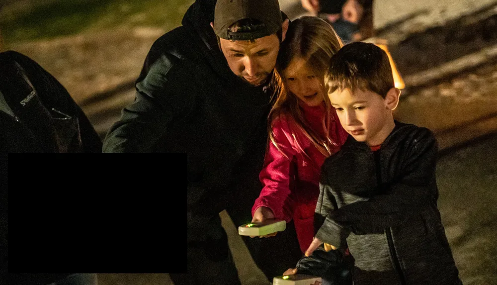An adult and two children seem engaged in an activity at night while holding what appears to be illuminated objects
