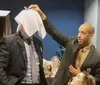 A man is playfully lifting a napkin over the head of another person whose head is not visible creating a humorous and candid moment in a social setting