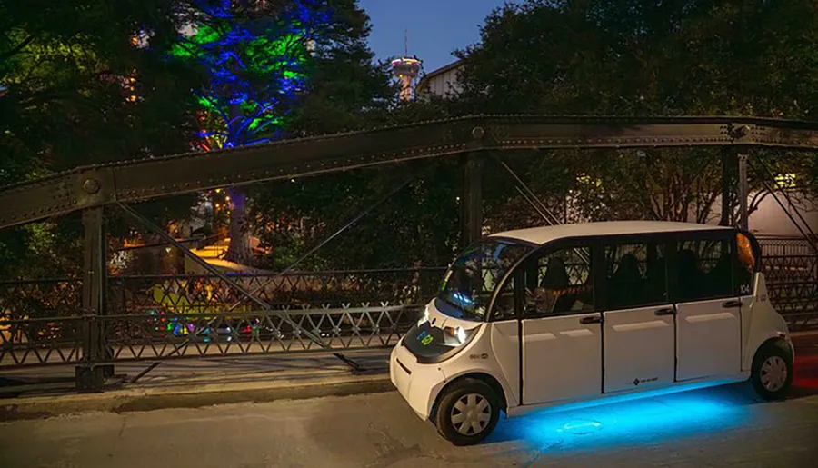 A compact electric vehicle with blue underglow lighting is parked next to an old truss bridge at dusk, with trees illuminated by colorful lights in the background.