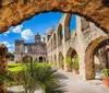 The image showcases the historic stone arches and architecture of an ancient mission against a backdrop of a bright blue sky with fluffy clouds