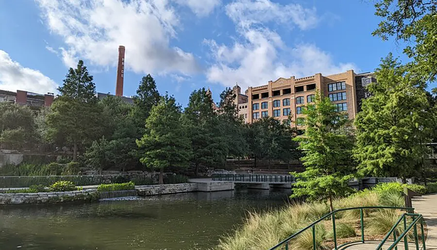 The image captures a serene urban park with a pond, green trees, a walking bridge, and historical industrial buildings in the background under a blue sky with scattered clouds.