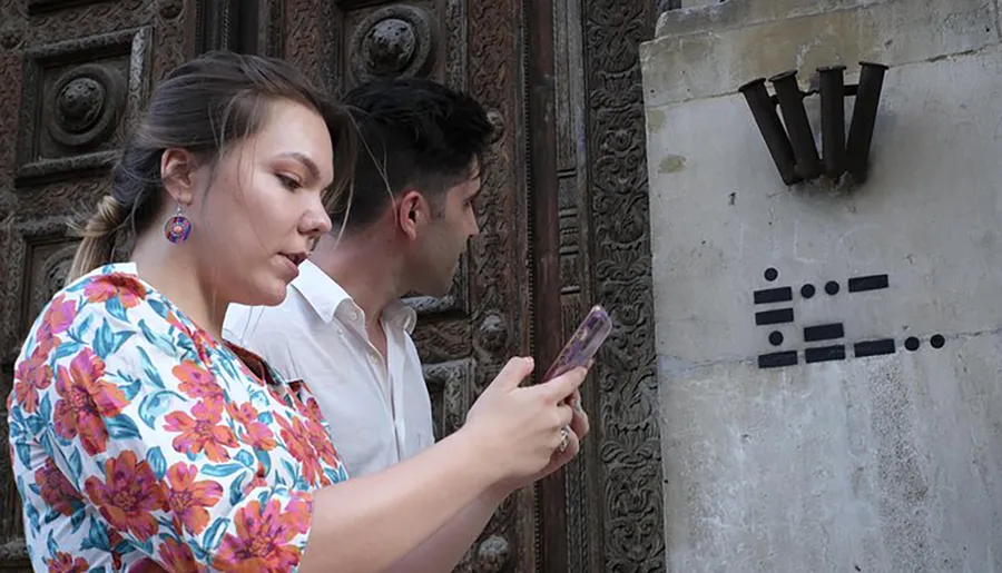 A woman is examining her phone while standing next to a man, with both of them seemingly ignoring or unaware of the artistic arrangement of metal objects on the wall beside them.