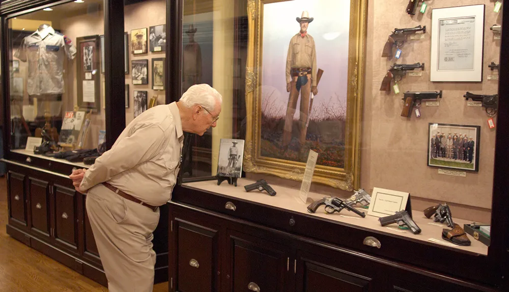 An elderly man is observing a museum display case containing firearms and memorabilia with a large portrait of a historical figure in the background