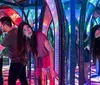 Several people navigate a colorful and reflective mirror maze with vibrant lights creating a kaleidoscopic effect