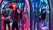 Several people navigate a colorful and reflective mirror maze, with vibrant lights creating a kaleidoscopic effect.