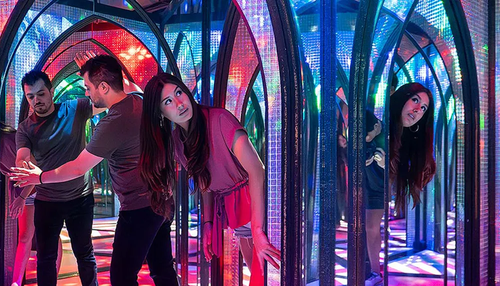 Several people navigate a colorful and reflective mirror maze with vibrant lights creating a kaleidoscopic effect