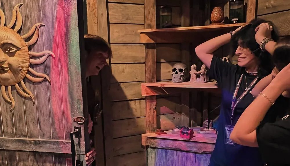 Two people react with surprise in a themed room with a wooden door and a sun sculpture as a third person peeks through the door