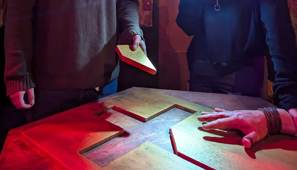 Two people are interacting with a wooden puzzle on a table one holding a piece while the other has a hand resting on the table illuminated by red lighting