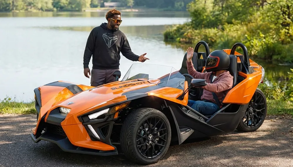 A person in a helmet sits in an orange three-wheeled vehicle while another person stands beside it near a scenic lake