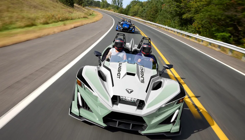 The image shows two Polaris Slingshot three-wheeled vehicles driving down a scenic road with passengers wearing helmets