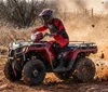 A person in full protective gear is riding a red ATV on a dirt trail kicking up dust and mud