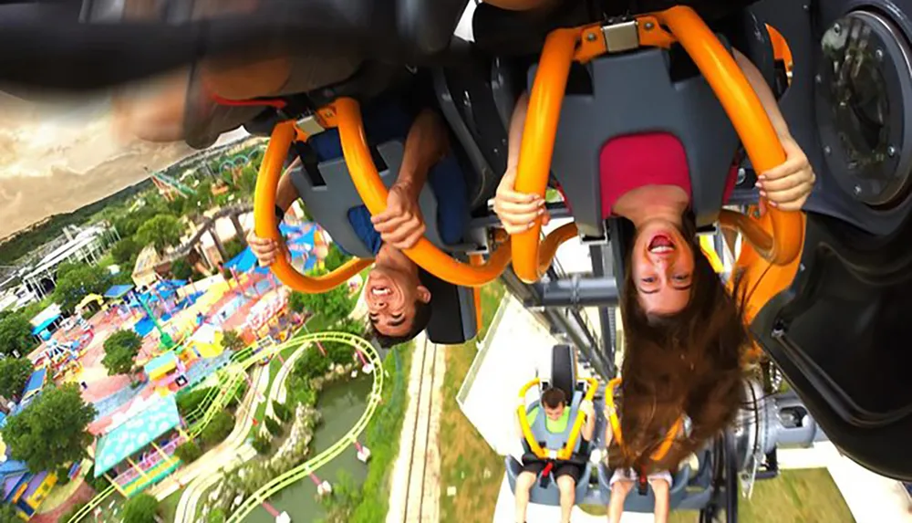 Two people are upside down on a roller coaster expressing excitement and thrill with an amusement park visible in the background