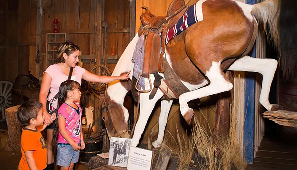 A woman and two children are interacting with a display of a horse in a museum-like setting