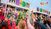 A festive Mardi Gras parade features colorful performers on a float, with a lively crowd reaching up towards them.