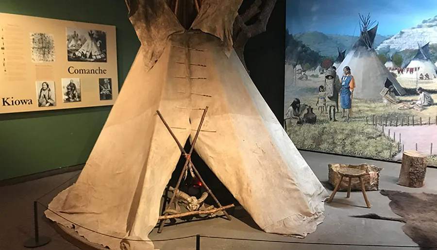 The image shows a museum exhibit featuring a tipi, artifacts, and informative displays about the Comanche and Kiowa peoples.