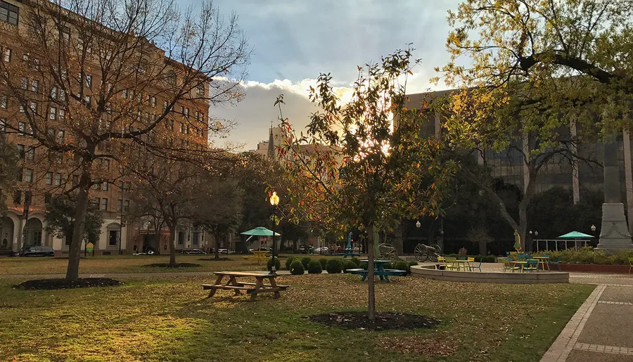 The image depicts a serene urban park with benches and a table, framed by autumnal trees against a backdrop of tall buildings, bathed in the soft glow of a setting sun.