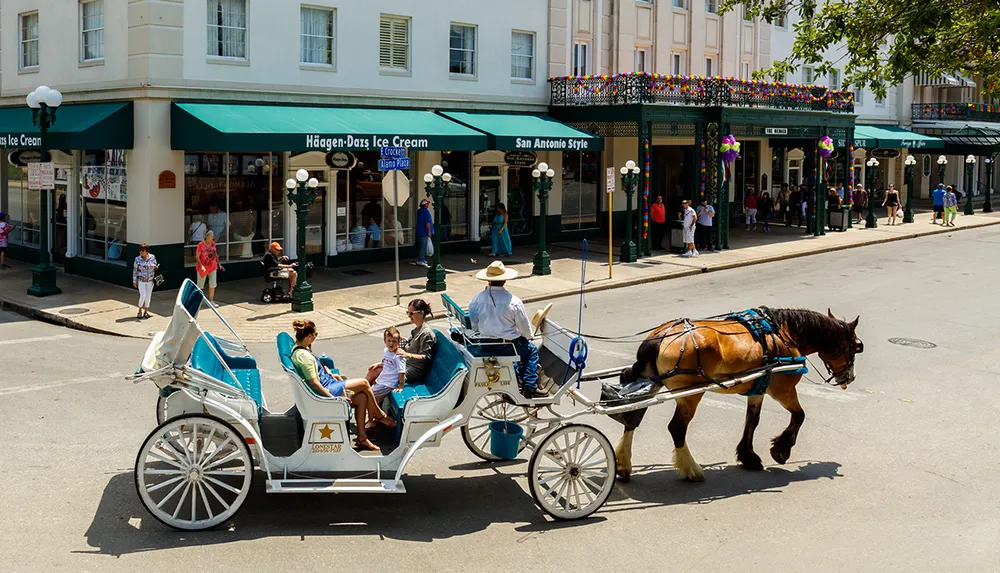 A horse-drawn carriage with passengers is being led by a driver wearing a cowboy hat on a sunny street with pedestrians and shops in the background