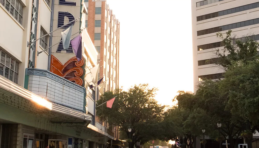 The image captures an urban street scene at sunset with the warm light of the setting sun filtering through trees highlighting a vintage-style theater marquee on the left and contemporary buildings in the background