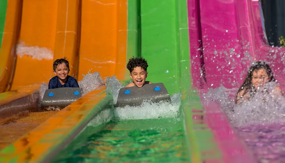 Three children express joy as they splash down colorful water slides on a bright sunny day