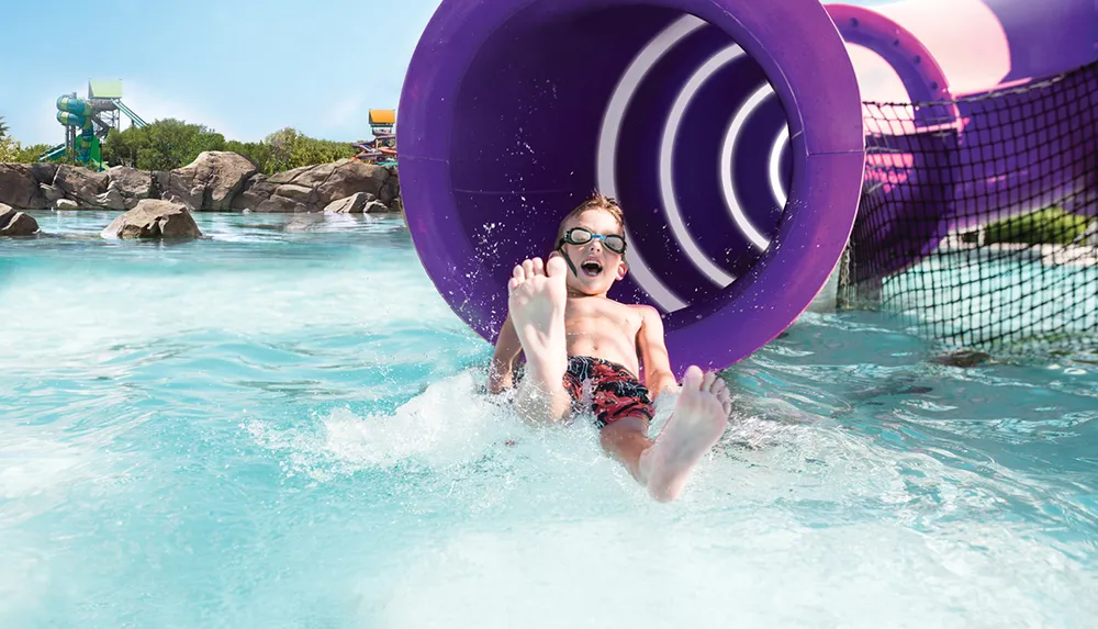 A child wearing goggles slides down a purple water slide with evident excitement