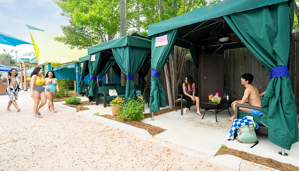 People are enjoying a day at a water park with some relaxing in cabanas and others walking by in swimwear