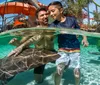 A man and a young boy are enjoying interacting with stingrays in a clear pool at a water park with slides and palm trees in the background
