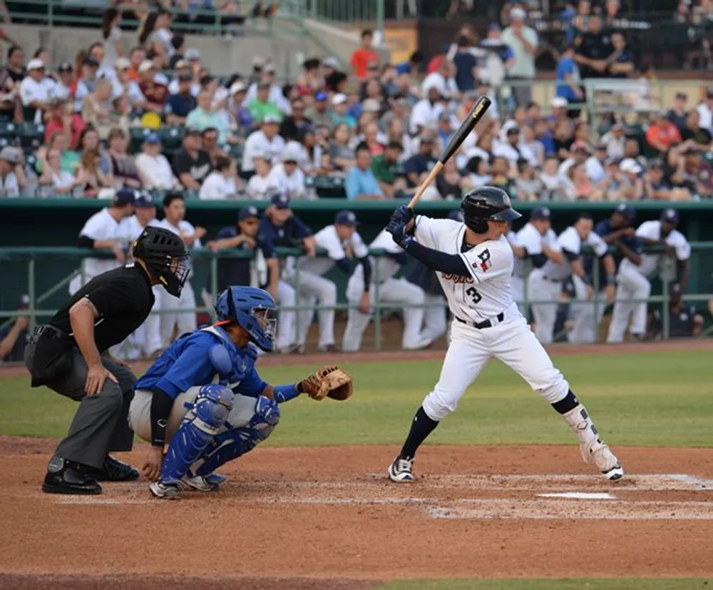 A baseball batter is in the midst of a swing as the catcher and umpire closely observe the play in a stadium filled with spectators