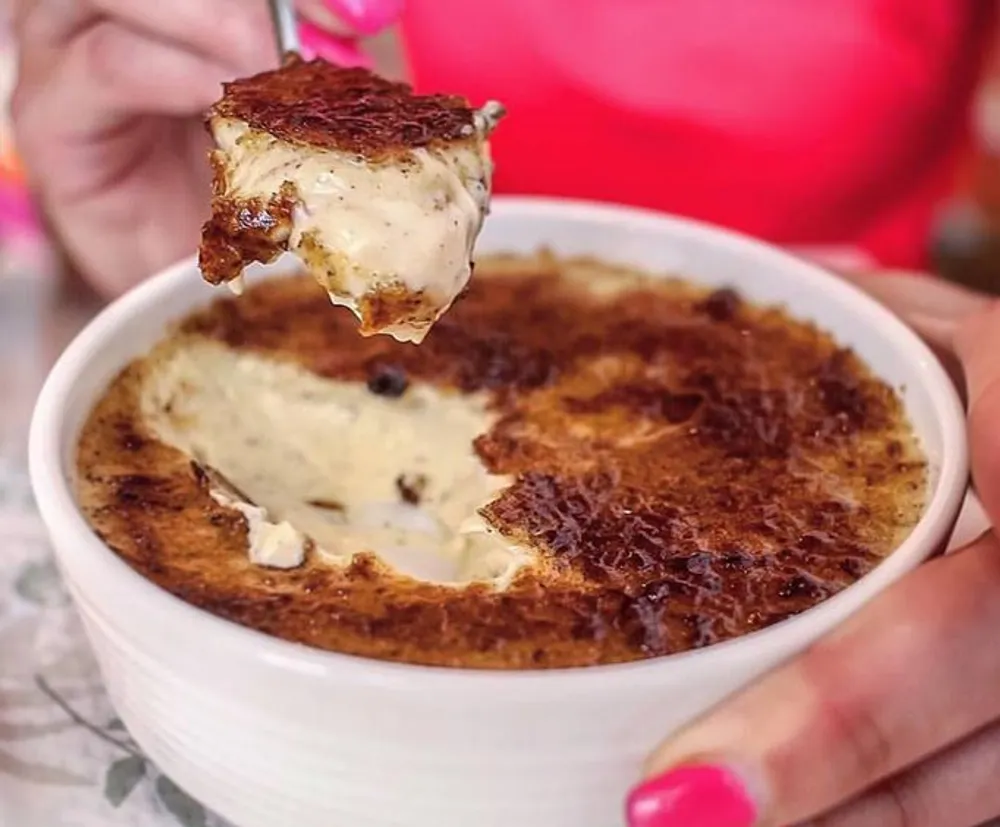 A person is holding a spoonful of creamy baked dip with a golden-brown crust taken from a white bowl