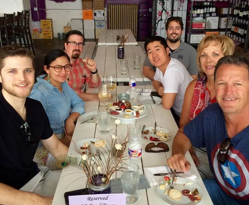 A group of seven people is smiling at the camera while sitting around a dining table with the remnants of a meal and drinks in a casual brightly lit setting