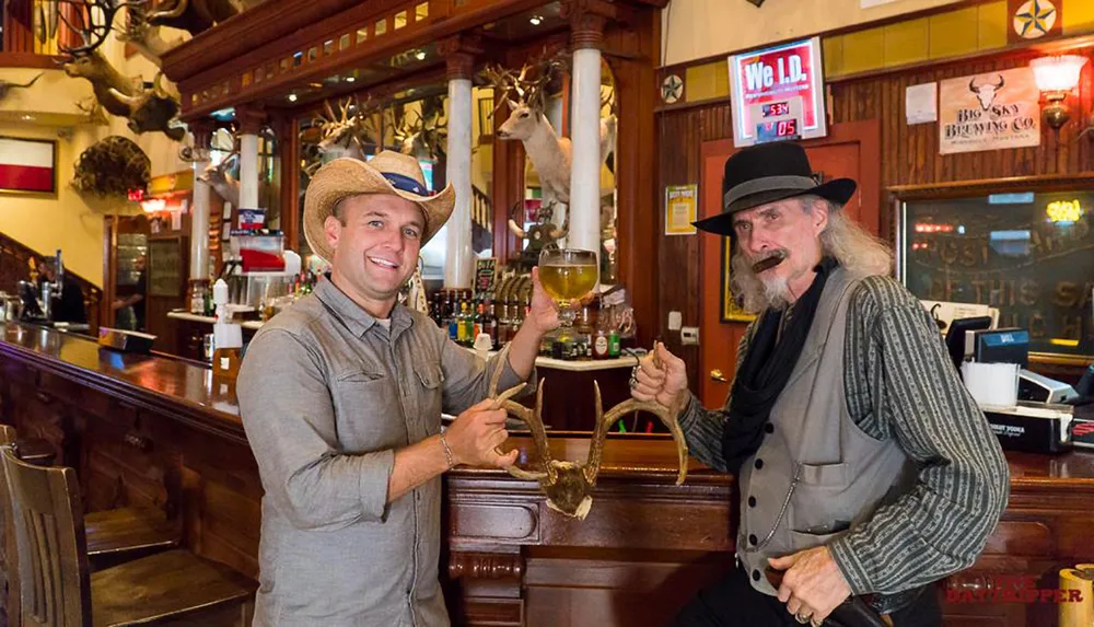 Two smiling men one wearing a cowboy hat and the other a top hat and with a handlebar mustache are toasting with a glass and antlers inside a bar decorated with taxidermied animals
