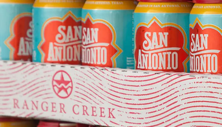 The image displays colorful cans with the label SAN ANTONIO behind a cardboard box marked RANGER CREEK, suggesting they are products, possibly beverages, from San Antonio, Texas.