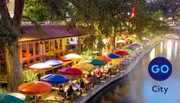 An outdoor riverside dining area illuminated at night with vibrant, colorful umbrellas and people enjoying their evening.